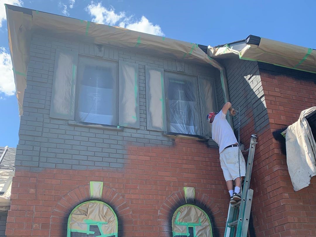 spray painter doing exterior painting on a large brick house, painting it from orange to grey.
